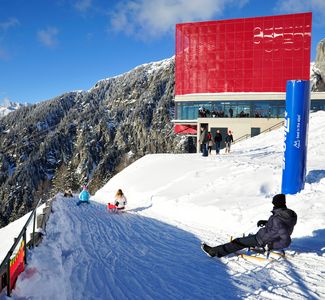 [Translate to englisch:] Menschen rodeln im Schnee an einer Bergstation mit rotem Gebäude in den Alpen, umgeben von verschneiten Bergen und klarem Himmel
