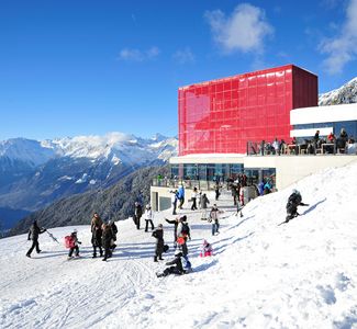 [Translate to englisch:] Menschen auf schneebedeckter Bergstation mit rotem Gebäude und verschneiten Bergen im Hintergrund