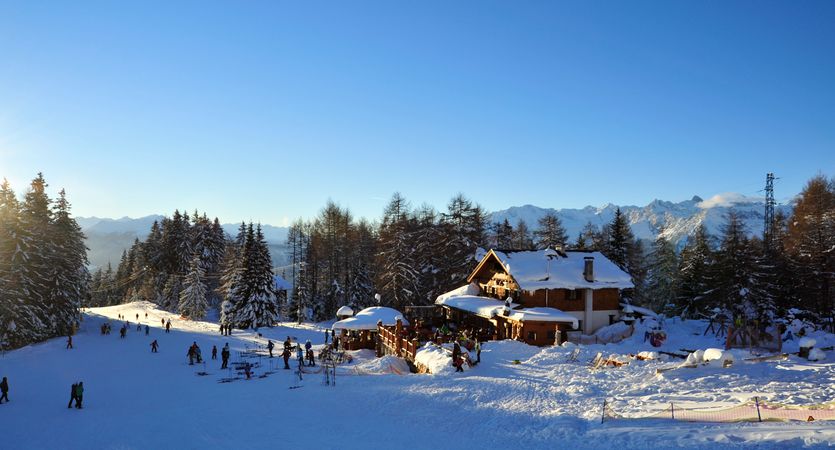[Translate to englisch:] Verschneite Skipiste mit Skifahrern, einer Berghütte und bewaldeten Bergen vor einem klaren blauen Himmel.