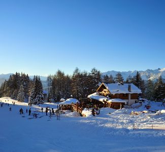 [Translate to englisch:] Sonnige Winterlandschaft mit verschneiter Berghütte und Menschen beim Skifahren umgeben von Tannenbäumen und Bergen