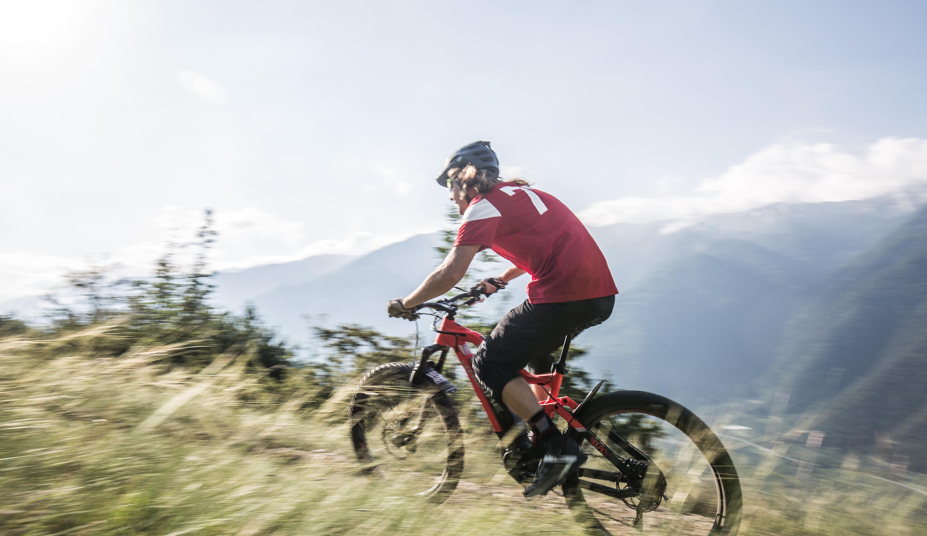 Mountainbiker in rotem Trikot fährt schnell einen grasigen Weg bergab mit Bergen im Hintergrund