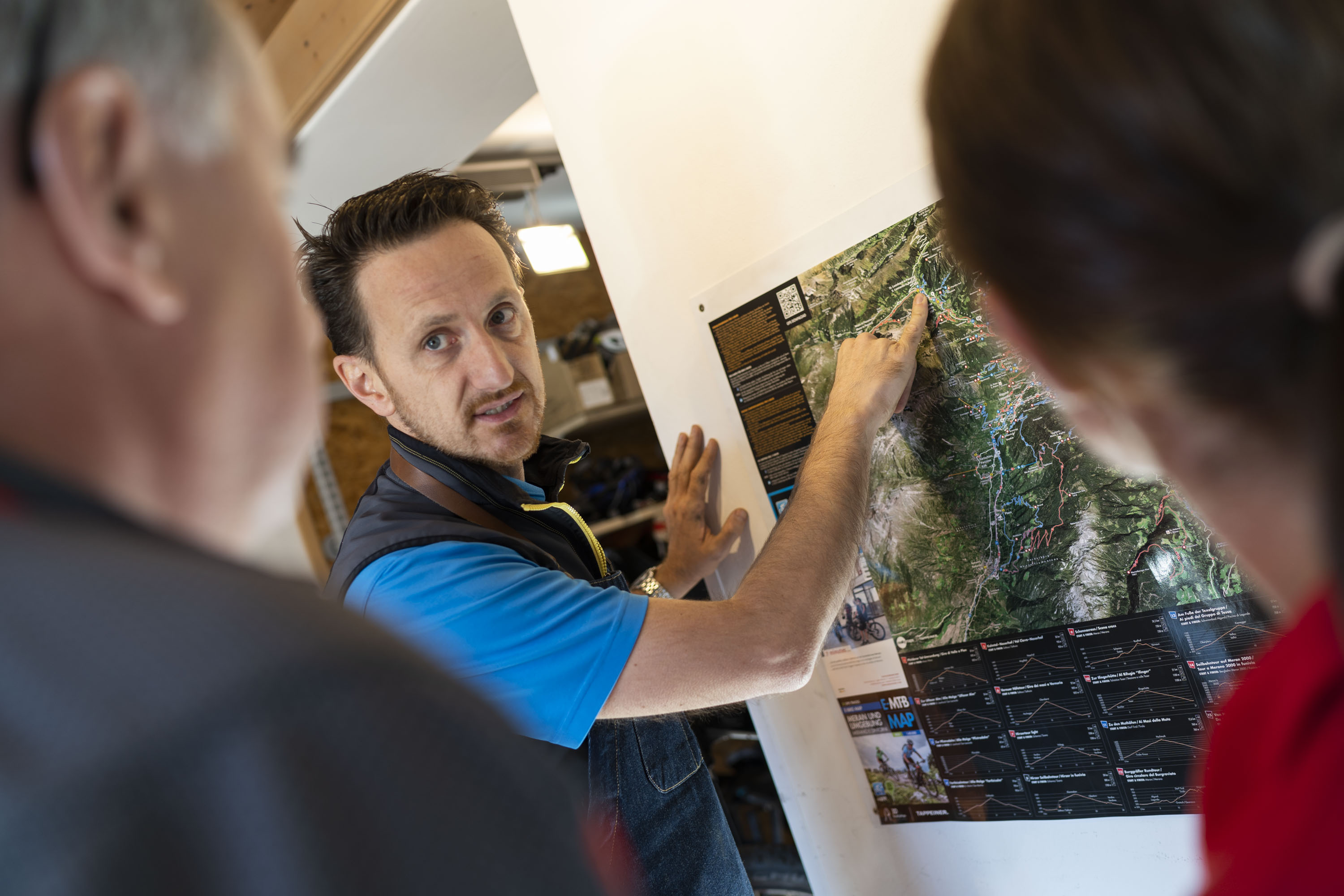 Ein Mann erklärt zwei Personen einen Wander- oder Radweg auf einer Karte an der Wand.