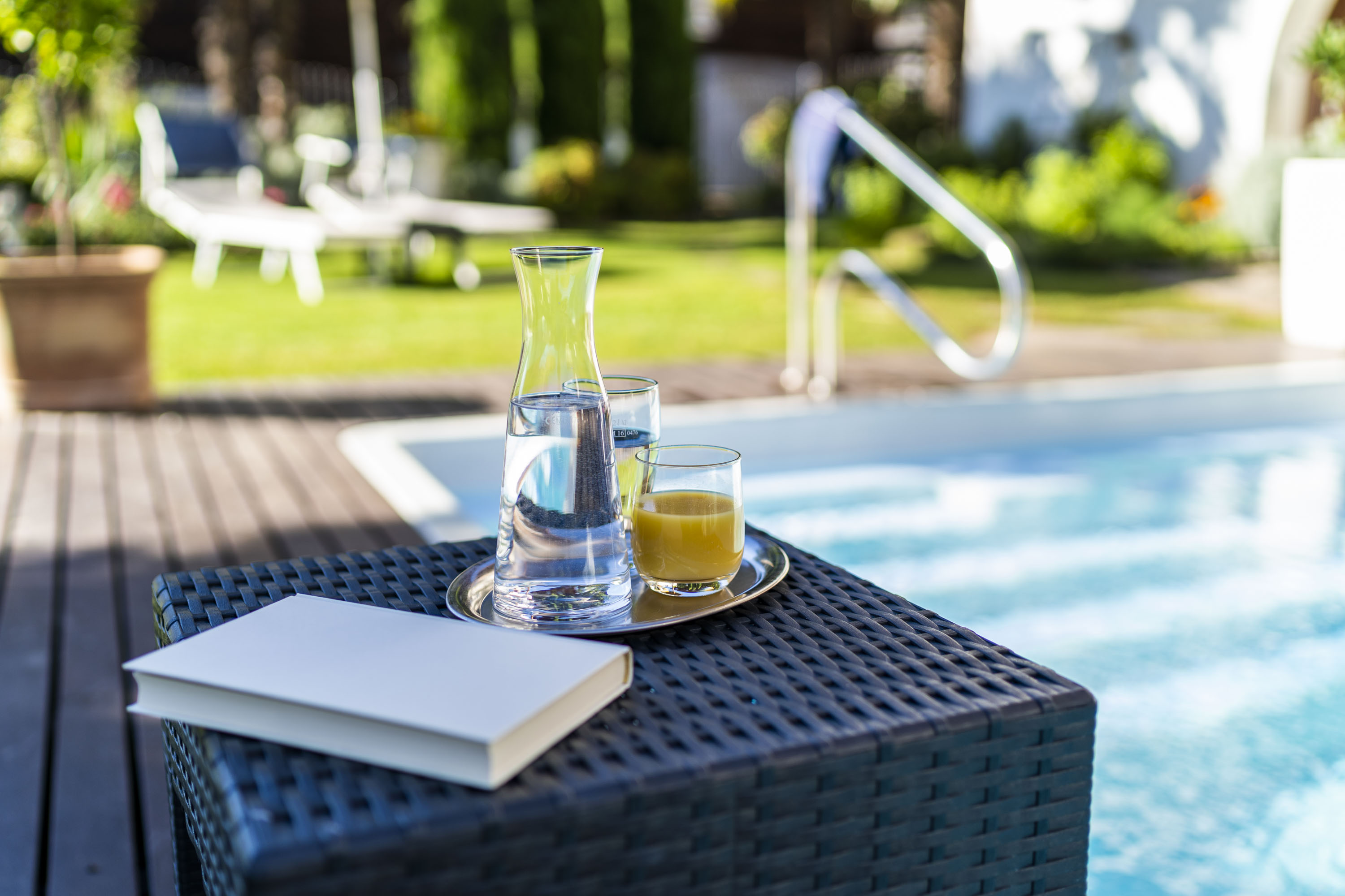 Tisch am Pool mit Karaffe Wasser, Glas Orangensaft und Buch, unscharfer Garten im Hintergrund