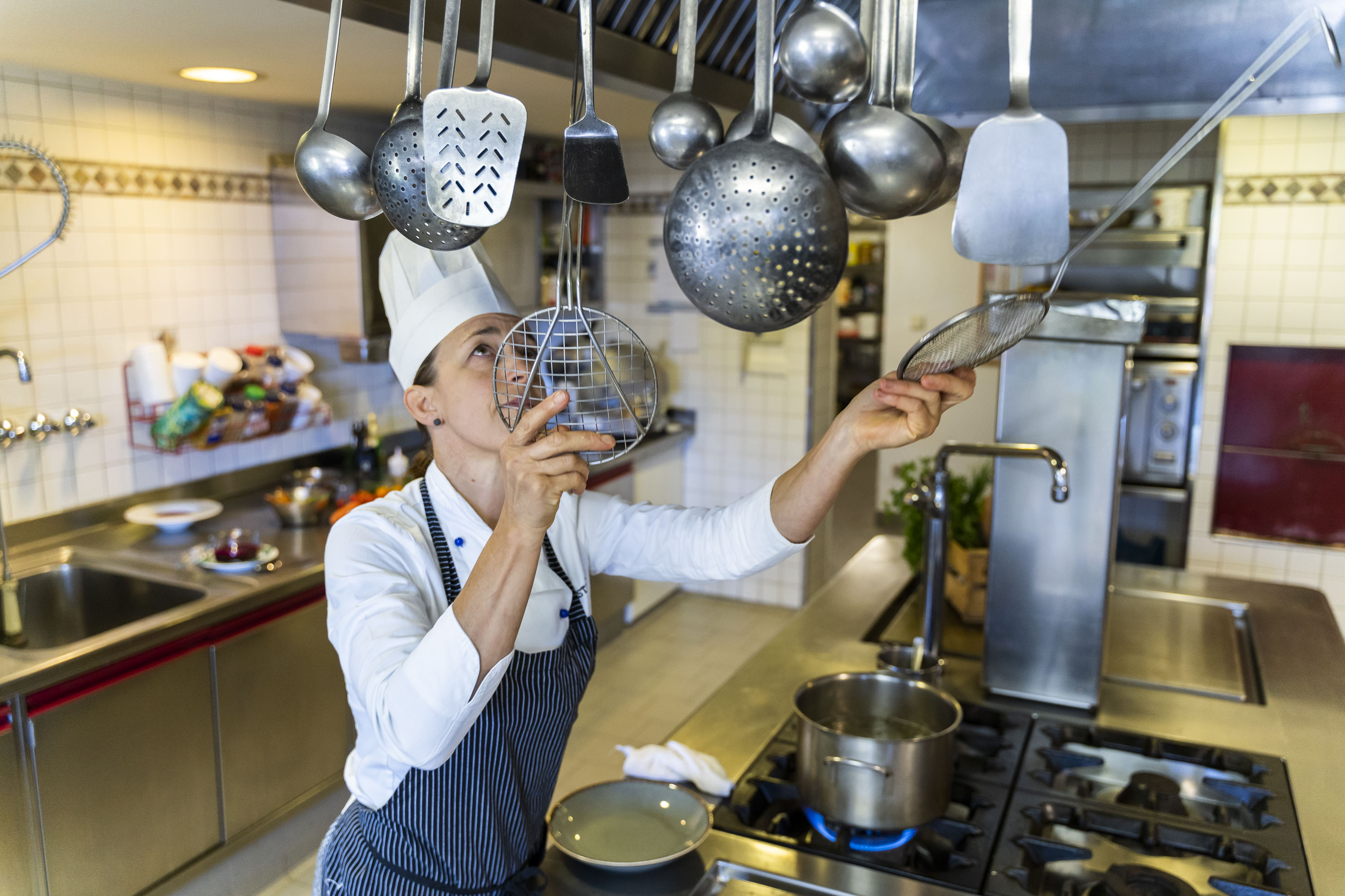 Küchenchef in weißer Uniform und Mütze greift in einer professionellen Küche nach hängenden Kochutensilien über einem Gasherd.