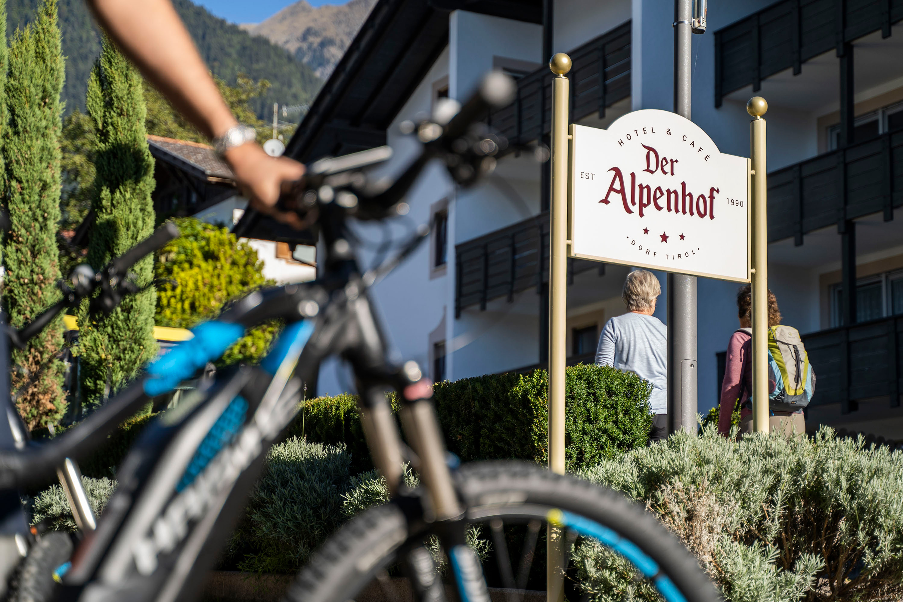 Fahrradfahrer im Vordergrund, Schild mit Hotelname Der Alpenhof, zwei Menschen gehen entlang eines Gebäudes in den Bergen.