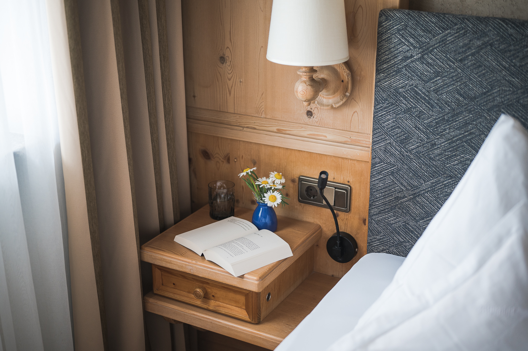 Nachttisch mit offenem Buch, Vase mit kleinen Blumen, Trinkglas, Leselampe und grauem gepolstertem Kopfteil eines Betts in einem hellen Raum mit Vorhängen