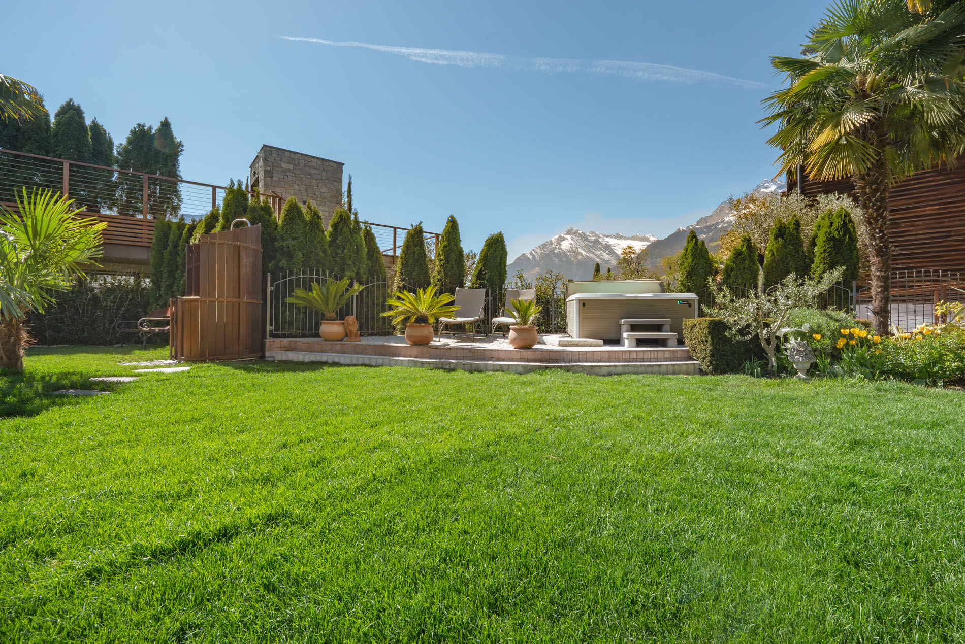 Grüner Garten mit Whirlpool, Liegestühlen und Blick auf schneebedeckte Berge bei klarem Himmel.