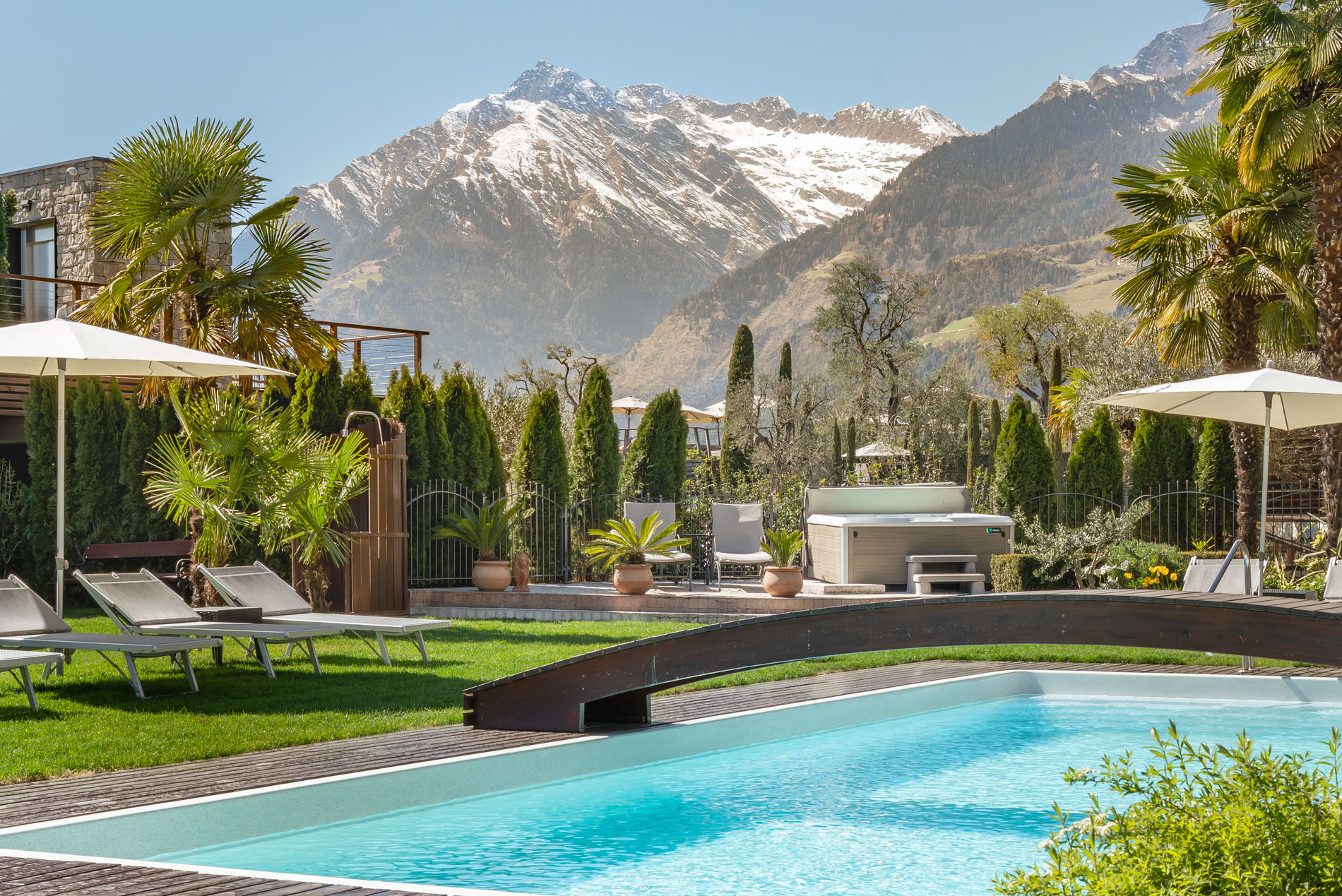 Outdoor-Pool mit Liegestühlen und Sonnenschirmen, umgeben von Palmen und Bergen mit Schneespitzen im Hintergrund.